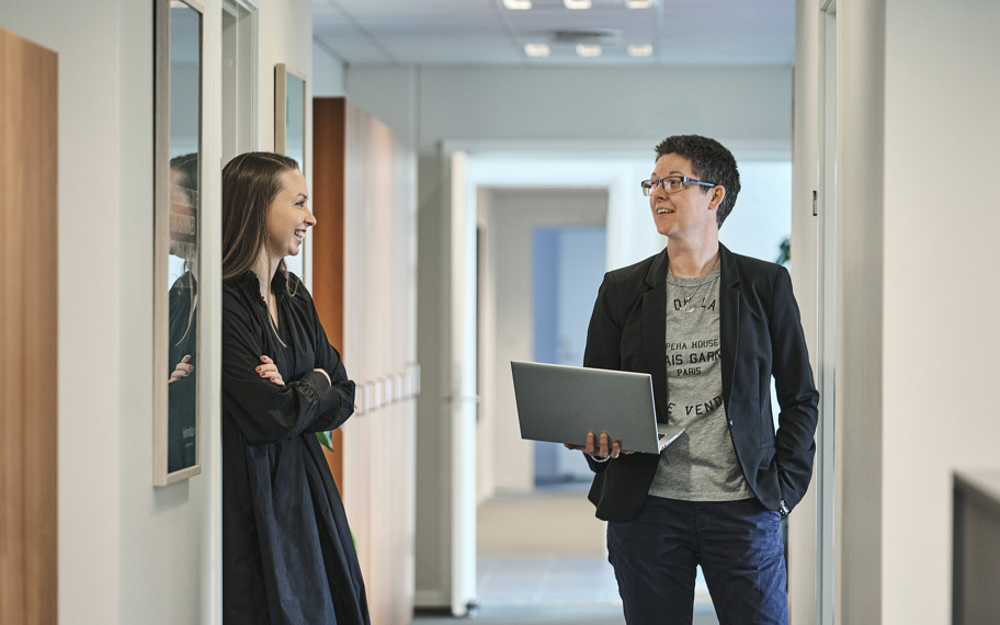 Ditte Marie Hansen og Jeanette Dich Sørensen med bærbar i hånden står og taler sammen på gang